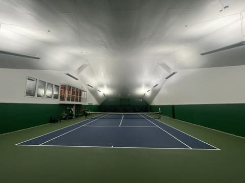 Court 2 seen from behind the baseline: a blue court with a wide green surround under a bright white ceiling, with the viewing-balcony windows high on the left wall.