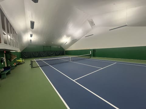 Court 2 from beside the net, with the balcony railing above the left side and ball baskets and benches along the wall.