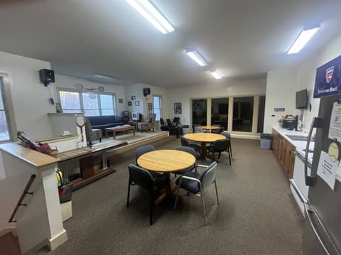 The kitchen and social lounge: round tables and chairs, a sofa area by the windows, and a kitchen counter with a refrigerator along the right wall.